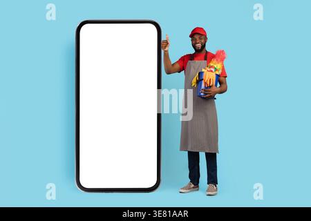 Schwarzer männlicher Reiniger mit Kappe und Schürze, die Daumen nach oben zeigen und den Korb mit Reinigungswerkzeugen neben dem großen Smartphone gegen blauen Hintergrund halten... Stockfoto