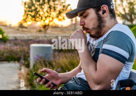 Arabischer Mann mit Kappe und Ohrhörern, der auf das Smartphone blickt, das am sonnigen Tag im Park sitzt Stockfoto