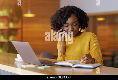 Schwarze Geschäftsfrau, die Notizen schreibt, während sie am Tisch mit Laptop im Café telefoniert Stockfoto