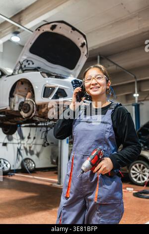 Chinesische Mechanikerin, die Overalls und Schutzbrille trägt, einen kabellosen Bohrer hält und einen Anruf hat, während sie wegblickt, während sie in der Nähe des Autos steht... Stockfoto