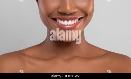 Zugeschnittenes Bild einer lächelnden schwarzen Frau, die ihre weißen Zähne vor grauem Hintergrund im Studio zeigt Stockfoto