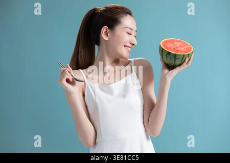 Fröhliche Chinesin in weißem Kleid, die Wassermelone hält, während sie im Studio vor blauem Hintergrund steht Stockfoto