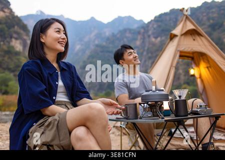 Ein lächelndes chinesisches Paar, das während des Campingens am Tisch sitzt und in der Nähe des Flusses und der Berge frühstückt Stockfoto