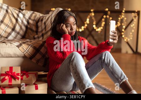 Verärgert Schwarze Teenager-Frau lehnt Gesicht auf geschlossene Faust beim Videoanruf mit dem Smartphone, das neben Weihnachtsgeschenkboxen auf dem Boden im lebenden Ro... Stockfoto
