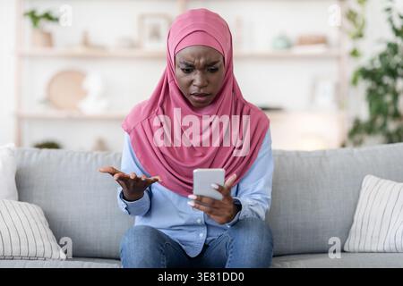 Verwirrte Schwarze Frau, die Hijab mit Smartphone trägt, während sie zu Hause auf dem Sofa sitzt Stockfoto
