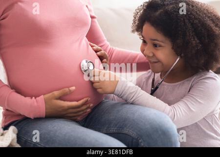 Schwarzes Mädchen, das ihren schwangeren motherÂ Bauch mit einem Stethoskop hört und Arzt zu Hause spielt Stockfoto