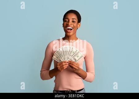 Schwarze Frau mit kurzem Afro, die Geld hält, während sie vor blauem Hintergrund im Studio steht Stockfoto