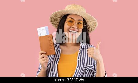 Fröhliche asiatische Frau, die Strohhut und Sonnenbrille trägt, einen Pass mit Flugtickets hält und die Daumen hoch zeigt, während sie gegen rosa Hintergras steht... Stockfoto