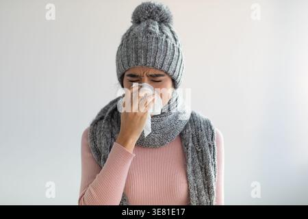 Kranke indische Frau, die Mütze und Schal trägt, die ihre Nase weht, während sie mit geschlossenen Augen vor grauem Hintergrund im Studio steht Stockfoto