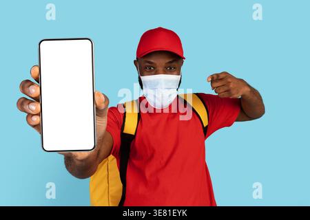 Schwarzer Liefermann mit Kappe und Gesichtsmaske, der Thermotasche trägt, zeigt und zeigt auf Smartphone mit leerem Bildschirm, der vor blauem Hintergrund steht... Stockfoto