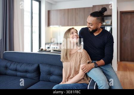 Fröhliches Nahost- und kaukasienpaar, das sich zuhause auf dem Sofa auf dem Sofa ansieht und miteinander spricht Stockfoto