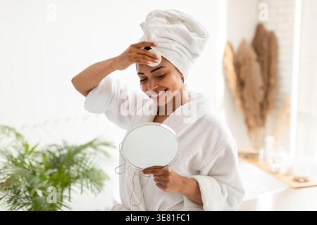 Schwarze Frau im Bademantel, die Haut pflegt und das Gesicht mit Wattepads abwischt, die in den Spiegel schauen, während sie zu Hause im Badezimmer steht Stockfoto