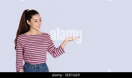 Lächelnde schwarze Frau in lässigem Outfit, die ihre offene Hand ansieht, während sie im Studio vor grauem Hintergrund steht Stockfoto