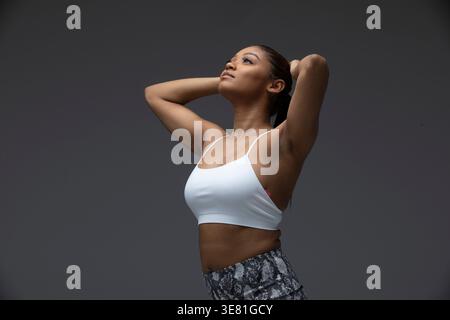 Schwarze Frau mit weißem Tanktop, die ihre Haare fixiert und im Studio vor grauem Hintergrund aufblickt Stockfoto