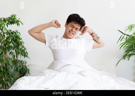 Selbstisolierung, guten Morgen, Faulheit und Aufwachen am Morgen am Wochenende. Glücklicher asiatischer Mann in weißem T-Shirt wacht auf, dehnt sich und sitzt auf dem Bett Stockfoto