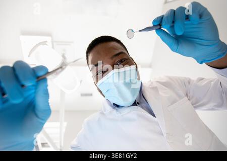 Schwarzer männlicher Zahnarzt mit Gesichtsmaske und Handschuhen, der Zahnwerkzeuge zur Untersuchung in der Klinik bereit hält Stockfoto