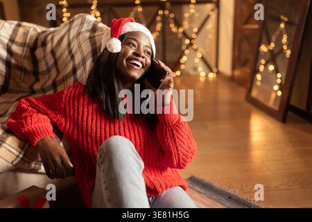 Schwarze Teenager-Frau, die Weihnachtsmann-Hut trägt und einen Telefonanruf gegen das Sofa hat, das zu Hause im Wohnzimmer auf dem Boden sitzt Stockfoto