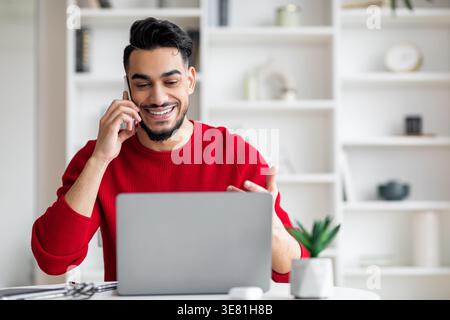 Ein arabischer Mann, der einen Anruf mit Gesten hat und auf den Laptop-Bildschirm schaut, der zu Hause am Tisch sitzt Stockfoto
