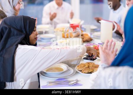 Lächelnde schwarze Frau im Hijab mit Freunden, die zusammen essen und zu Hause am Tisch sitzen Stockfoto