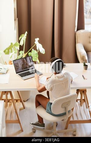 Rückansicht von Black Girl, das Kopfhörer trägt und beim Lernen Online-Unterricht auf einem Laptop im Esszimmer zu Hause ansieht Stockfoto
