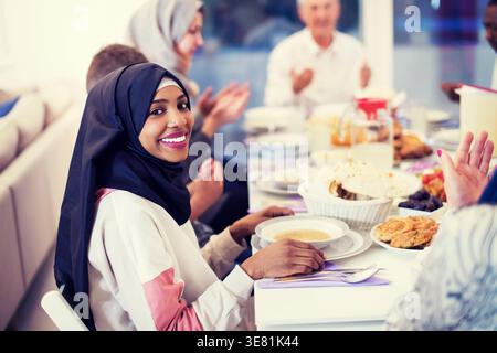 Lächelnde schwarze Frau im Hijab mit Familie und Freunden, die zusammen essen und zu Hause am Tisch sitzen Stockfoto