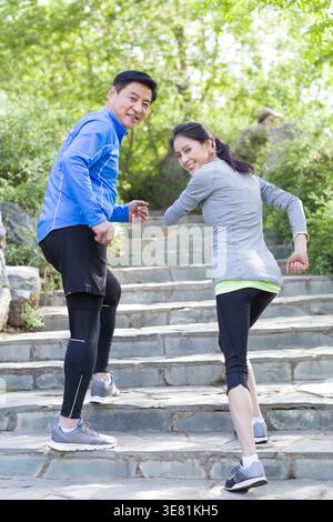 Lächelndes chinesisches Paar mittleren Alters in Fitnesskleidung, das tagsüber gemeinsam auf Treppen im Park läuft Stockfoto
