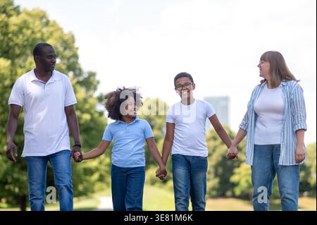 Schwarzer Mann, der die Hände mit Kindern und einer kaukasischen Frau hält, während er tagsüber zusammen im Park spaziert Stockfoto