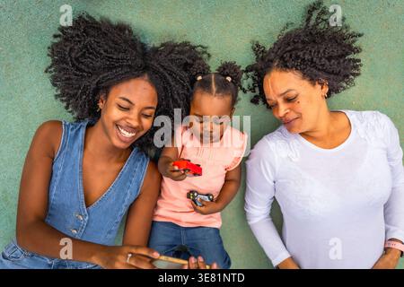 Schwarze Frau beobachtet auf dem Smartphone mit Mutter und Tochter, die auf dem Boden im Park liegen Stockfoto
