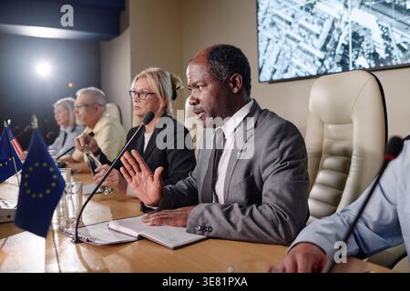 Schwarzer Politiker mittleren Alters, der im Mikrofon spricht und mit einem Journalisten spricht, während er während der Konferenz mit Kollegen am Tisch sitzt Stockfoto