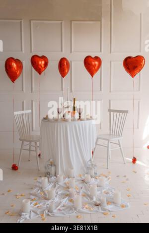 Romantisches Abendessen für zwei Personen mit roten Herzballons, weißer Tischdecke, Kerzen, Champagner und Rosenblättern in einem hellen, minimalistischen Zimmer zum Valentinstag Stockfoto