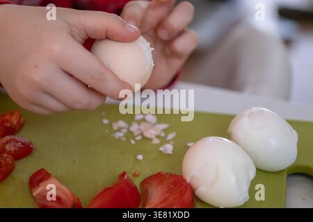 Nahaufnahme von Kinderhänden, die ein gekochtes Ei auf einem Schneidebrett mit Tomaten schälen. Ideal für Familienkochen, das Erlernen von Lebensmittelkenntnissen und die Entwicklung von Kindern Stockfoto