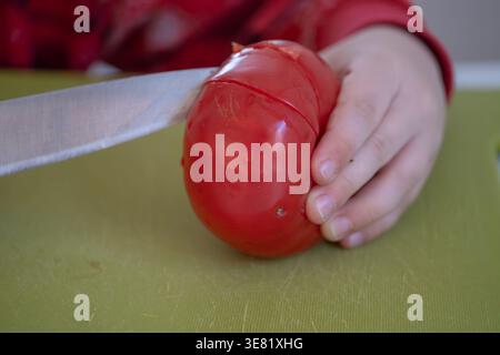 Nahaufnahme von Kinderhänden, die eine Tomate mit einem Messer auf einem Schneidebrett schneiden. Konzentrieren Sie sich auf das Erlernen grundlegender Kochfähigkeiten und die Zubereitung von Speisen. Ideal für Familien Stockfoto