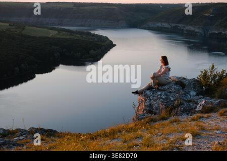 Eine junge Wanderer sitzt an einer felsigen Klippe und genießt bei Sonnenuntergang den malerischen Blick auf den Fluss Canyon. Friedliche Reisende, die sich während der goldenen Stunde in der Natur ausruhen Stockfoto
