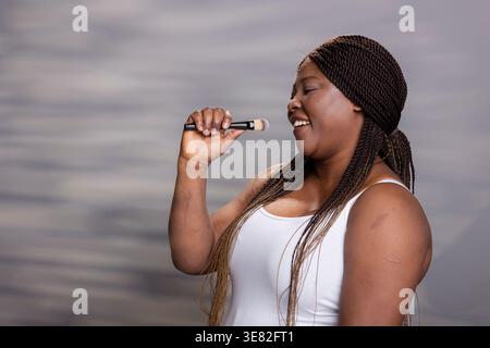 Alberne afroamerikanische Frau, die sich selbst vertraut und am Make-up-Pinsel singt, Spaß hat und durch natürlichen Make-up-Look Selbstliebe repräsentiert. Lustige Frau mit strahlender taufender Haut. Stockfoto