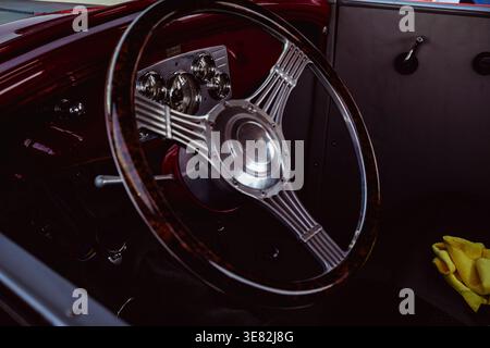 Das Vintage-Lenkrad glänzt in einem tiefroten Fahrzeuginnenraum. Chromspeichen reflektieren das Umgebungslicht elegant. Dunkle Holzverkleidungen betonen das klassische Design des Armaturenbretts Stockfoto