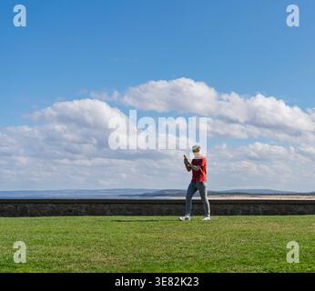 Ein Mann läuft über eine grüne Grasfläche, hält ein Telefon und trägt eine Sonnenbrille an einem hellen Tag in Lossiemouth, Moray, Schottland. Stockfoto