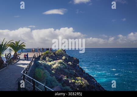 Eine kleine Gruppe von Touristen steht auf einem gepflasterten Klippen-Mirador über dem Atlantischen Ozean in Puerto de Santiago, eingerahmt von Kanarischen Palmen, Agaven und Sukkulentensträuchern am Eingang zu den Acantilados de los Gigantes an der Südwestküste Teneriffas auf den Kanarischen Inseln, Spanien. Die Wintersonne des Mittagnachmittags beleuchtet das Basaltkap und das tiefblaue Meer, fotografiert am 17. Januar 2026. Stockfoto