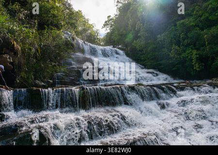 Das Wasser stürzt kraftvoll über eine Reihe von felsigen Felsvorsprüngen und schafft ein faszinierendes Naturspektakel innerhalb eines lebendigen grünen Waldes, mit einer Person, die sich erkundet Stockfoto