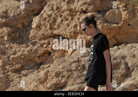 Eine junge Frau mit Sonnenbrille spaziert nachdenklich auf felsigem Boden in einer Wüstenumgebung und genießt einen warmen Tag inmitten natürlicher Schönheit. Stockfoto