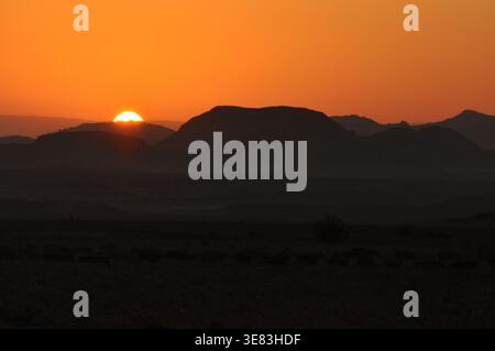 Die goldene Sonne untergeht hinter Silhouettenbergen in einer friedlichen Wüstenlandschaft. Das warme Leuchten erfüllt den Himmel und markiert den Übergang von Tag zu Nacht. Stockfoto