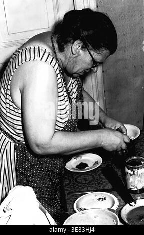 Slaviansk, 1950er Jahre Eine intime Szene, in der Nadezhda Juryjewna Kravelis alltägliche kulinarische Aufgaben erledigt. Sie wird gefangen, indem sie sich weit nach vorne über einen Küchentisch beugt und sich intensiv auf die Zubereitung von Speisen in der Nähe von Glasgläsern und Keramikplatten konzentriert. Mit einem gestreiften Oberteil, einer gemusterten Schürze und einer runden Brille verkörpert sie den fleißigen Geist der Epoche. Dieses authentische Vintage-Foto zeigt traditionelle Haushaltsroutinen und die anhaltende, warme familiäre Atmosphäre des häuslichen Lebens im friedlichen Donbass. Stockfoto