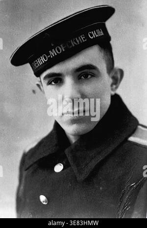 Slaviansk, 1950er Jahre Ein einzigartiges Studio-Porträt von Michail Iljitsch Kravelis, der spielerisch eine dunkle Marinekappe trägt, die mit der Inschrift „Naval Forces“ in kyrillischer Schrift versehen ist, geliehen von seinem Freund, dem zukünftigen Kapitän Anatoly Lamekhov, für dieses unvergessliche Foto. In einem schweren, dunklen Mantel mit Kragen gekleidet, blickt Mikhail unaufhörlich in die Kamera. Dieses offen inszenierte Schwarzweiß-Bild zeigt die junge Kameradschaft, persönliche Mode aus der Mitte des Jahrhunderts, starke Freundschaften und den warmen, optimistischen Geist, der das tägliche Leben im friedlichen Donbass definiert. Stockfoto