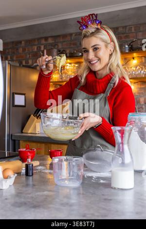 Frau trägt roten Pullover und graue Schürze mischender Teig in der Wohnküche mit Glasmischschschüssel Stockfoto