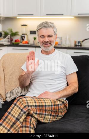 Ein Mann mittleren Alters zu Hause winkt zur Kamera, sagt Hallo und beginnt mit einem freundlichen Lächeln Online-Kontakt. Reifer Mann auf dem Sofa schaut auf die Linse nickt spricht kurze Begrüßung warme Stimmung offener Ton ruhiger Atem ruhig. Stockfoto