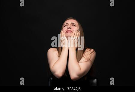Eine junge Frau, die ihr Gesicht bedeckt, während sie weint, Traurigkeit, Trauer und emotionalen Schmerz vor dunklem Hintergrund erlebt Stockfoto
