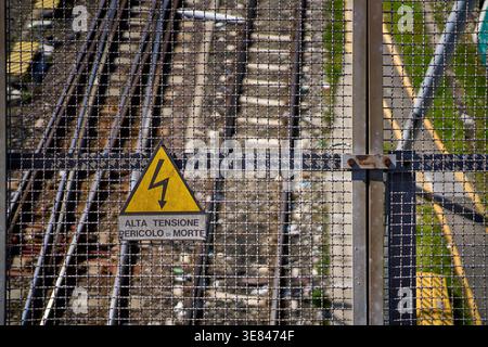 Nahaufnahme eines Hochspannungs-Warnschildes an einem Sicherheitszaun aus Drahtgeflecht am Bahnhof Bologna Centrale, Bologna, Emilia-Romagna, Italien. Stockfoto