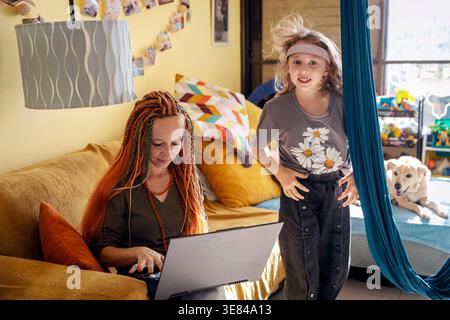 Mutter arbeitet an einem Laptop, während ihre aktive Tochter in der Nähe in ein Wohnzimmer springt. Berufstätige Mutter Stockfoto