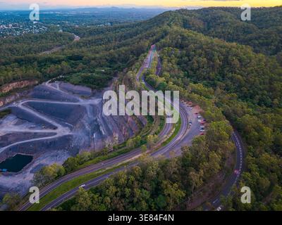 Aus der Vogelperspektive wird eine gewundene Straße und ein aktiver Steinbruch in einen dicht bewaldeten Hügel mit Blick auf die Stadt Brisbane während der Dämmerung erfasst. Stockfoto