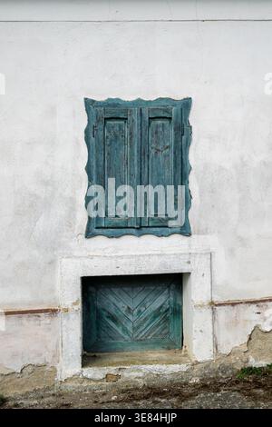 Die verwitterte Fassade eines alten Gebäudes verfügt über ein geschlossenes, blaublaues Fenster und eine passende Tür darunter. Die abblätternde Farbe und der rissige Putz suggerieren Stockfoto