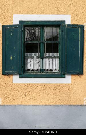 Ein Fenster mit dunkelgrünen Fensterläden und weißen Spitzenvorhängen wird von einer strukturierten gelben Wand eingerahmt. Die Fensterläden sind teilweise geöffnet, wodurch der Innenraum sichtbar wird Stockfoto